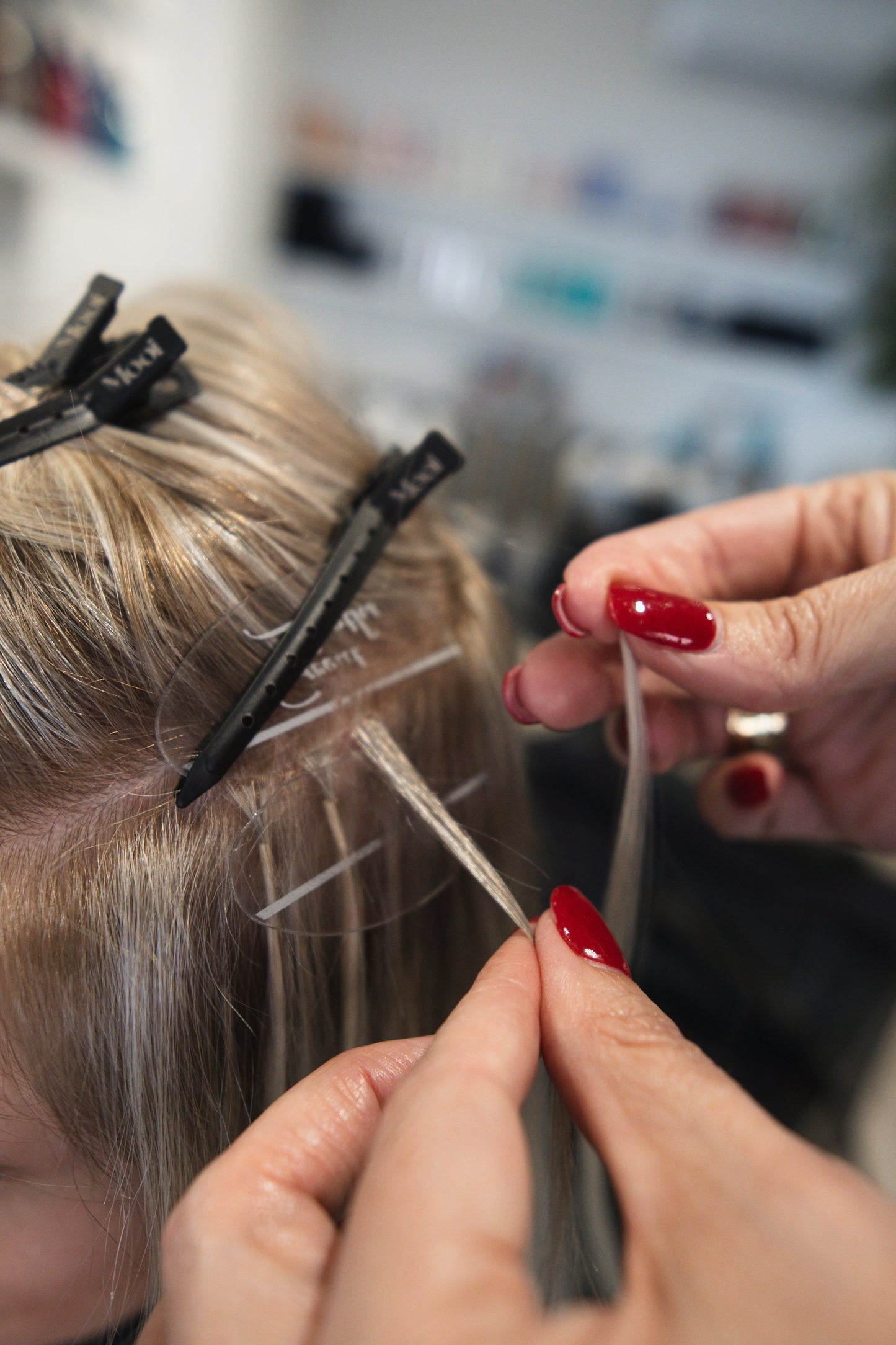 Close-up of a hairdresser's hands skillfully applying Mooi Hair’s Bonds Masterclass clip-in extensions to blonde hair, capturing hands-on training in the art of transformation. The nails are painted red, with the background blurred to emphasize this crucial stage of the hair extension journey.