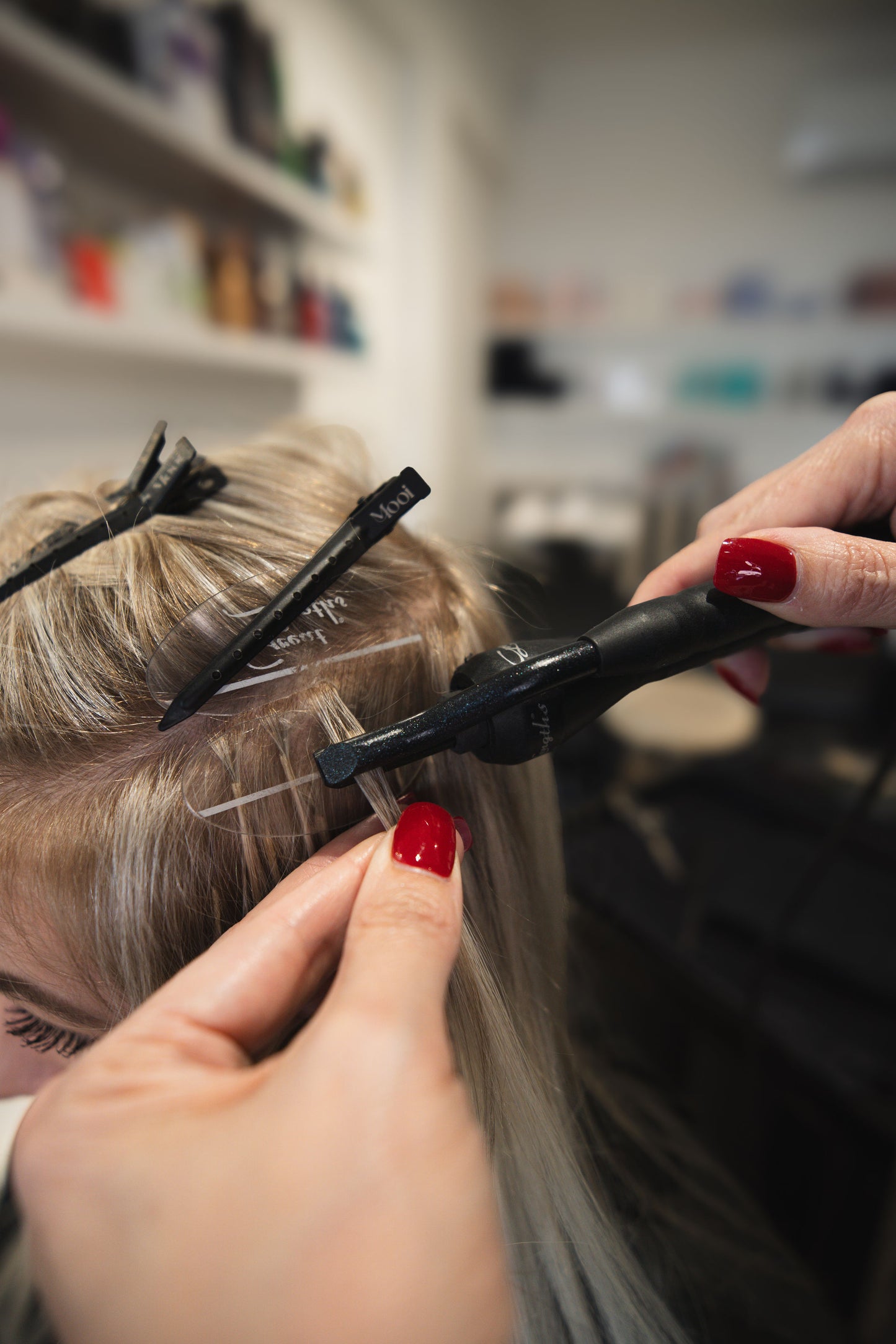 A person with long blonde hair is on a Bonds Masterclass journey by Mooi Hair, getting extensions applied. Their hair is sectioned and secured with clips while a stylist uses precision tools. Shelves filled with various Mooi Hair products are blurred in the background.