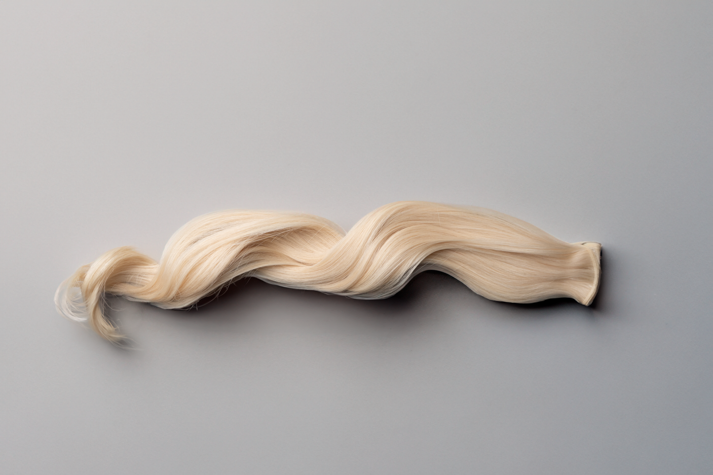 A single lock of long, wavy platinum blonde hair against a smooth, light gray background showcases the elegance of the Mooi Hair Seamless Weft | 50cm/20 inch extensions.
