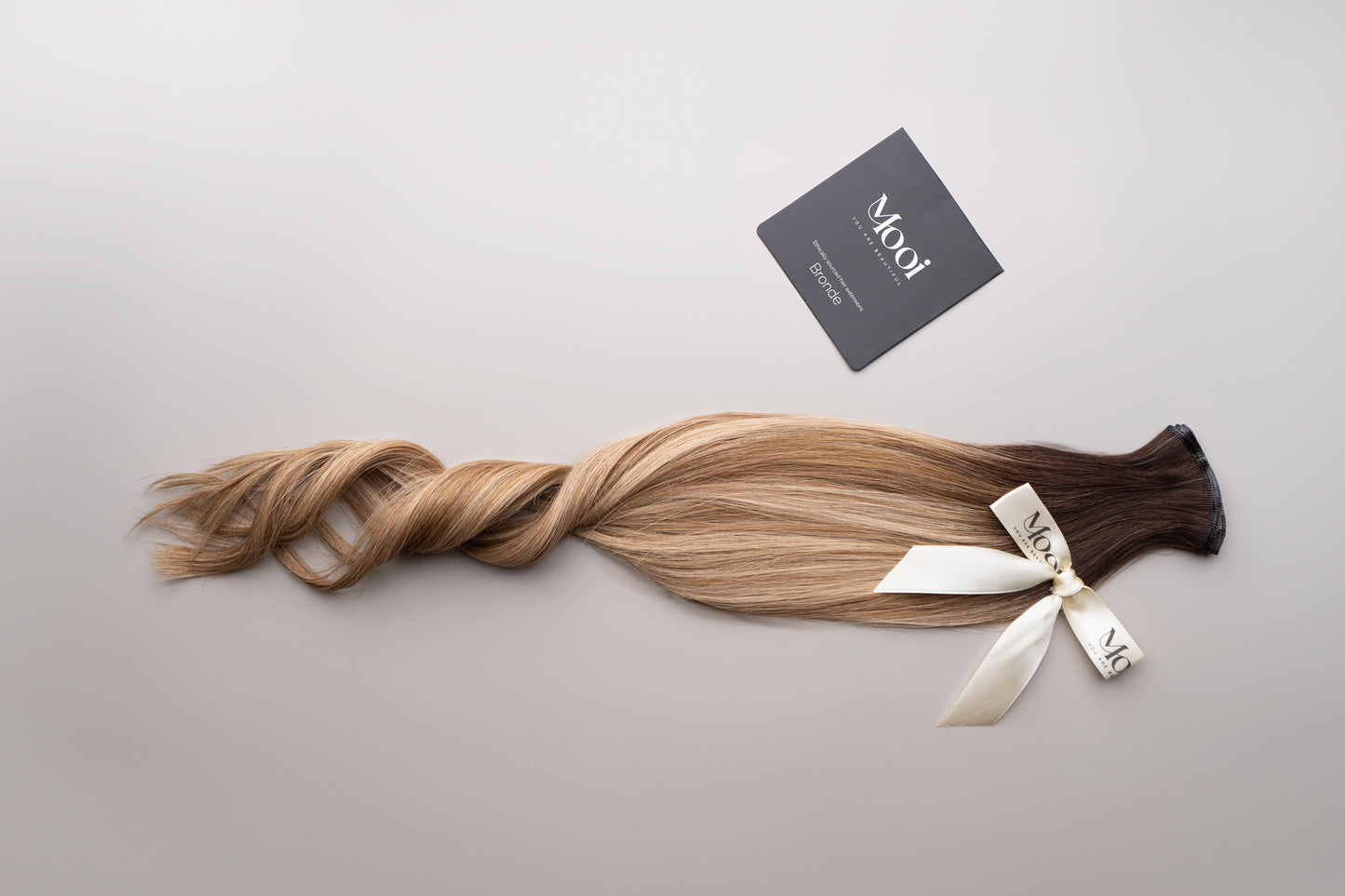 A long strand of the Seamless Weft in brown to blonde ombre from Mooi Hair is displayed on a light surface, elegantly tied with a cream ribbon. A black card featuring the "Mooi Hair" logo and additional details is nearby, suggesting that these 50cm/20 inch extensions can be customized to suit any style.