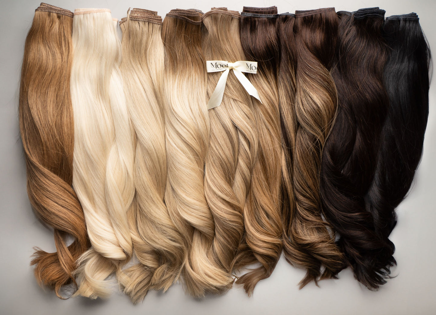 A lineup of Bonds Masterclass hair extensions by Mooi Hair, showcasing a spectrum of shades from light blonde to dark brown and black. Each wavy and glossy piece narrates its own hair extension story, with one adorned with a ribbon labeled "Moresoo.