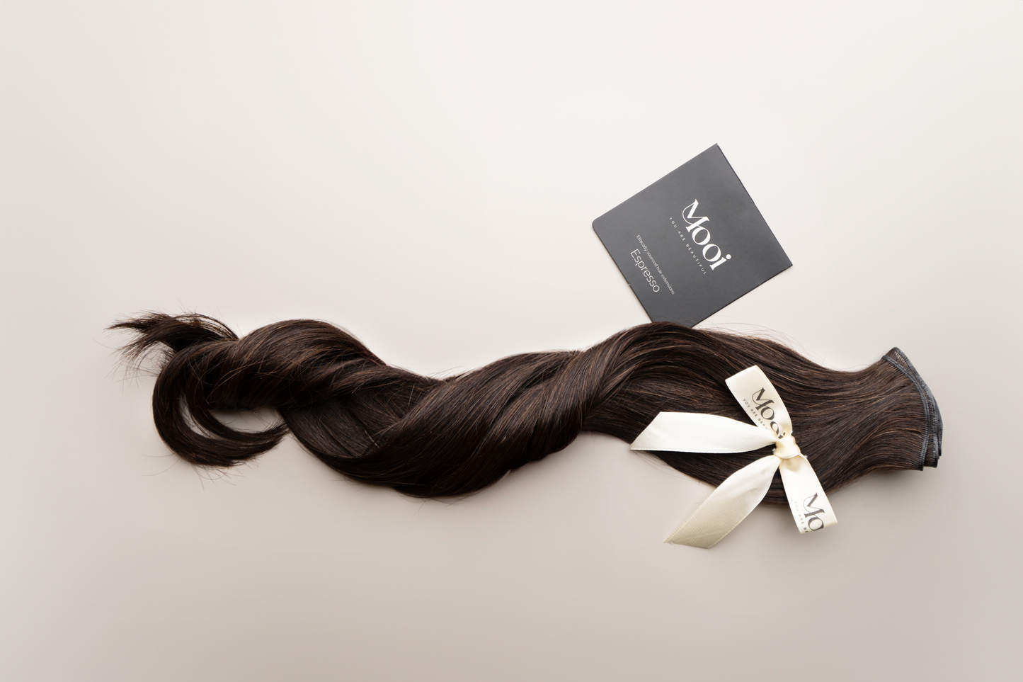 A bundle of long, dark brown Seamless Weft hair extensions tied with a cream-colored ribbon rests on a light surface. These 50cm/20 inch extensions feature a black square tag displaying the brand name "Mooi Hair," ensuring a customized fit for optimal comfort.