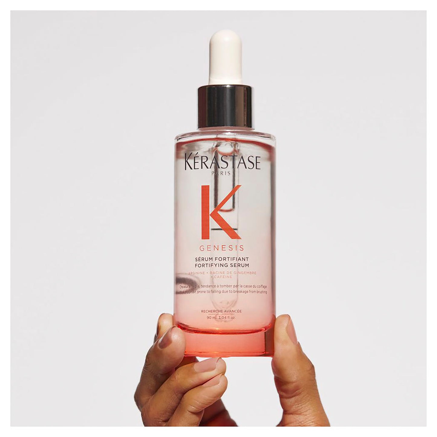 A person holds a bottle of Kerestase Genesis Homme Serum Anti-Chute Fortifiant 90ml, elegantly set against a plain white background. The transparent bottle with its subtle pink tint features a sleek white dropper cap and an eye-catching label in black and pink text, embodying luxury and care in every drop.