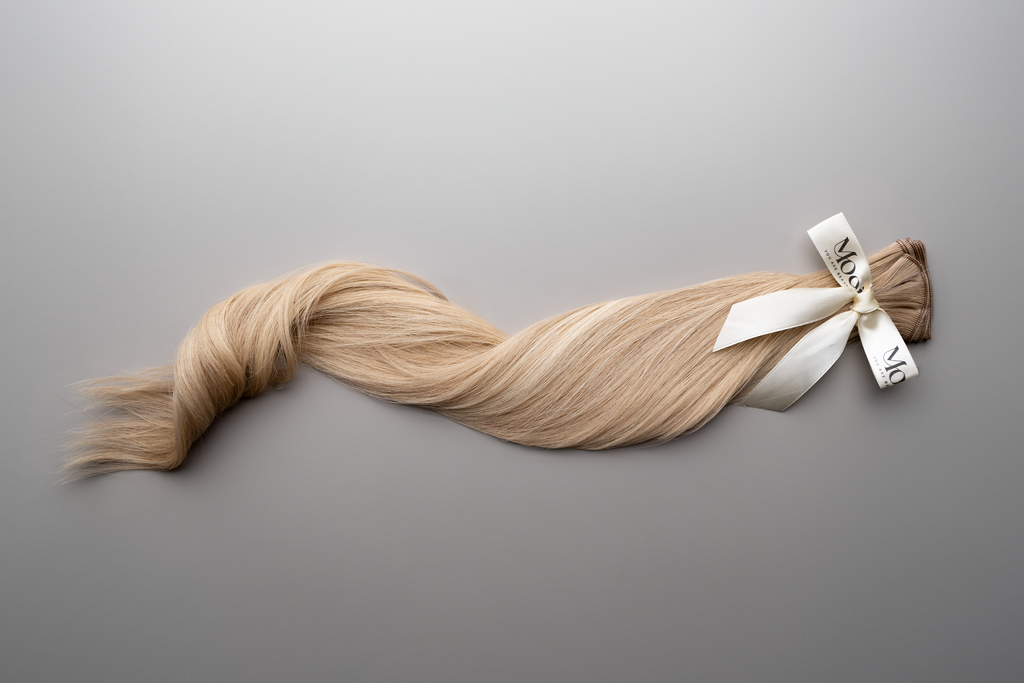 A bundle of long, smooth, blonde hair tied with a white ribbon on a gray background highlights the exquisite charm of Mooi Hair's Seamless Weft | 50cm/20 inch extensions.