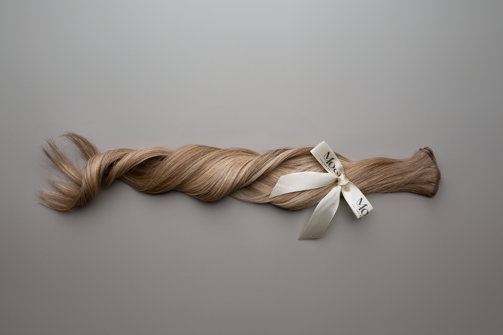 A long, braided lock of light brown hair tied with a white ribbon, laid on a smooth gray surface, mirrors the elegance of Mooi Hair's Seamless Weft | 50cm/20 inch extensions.