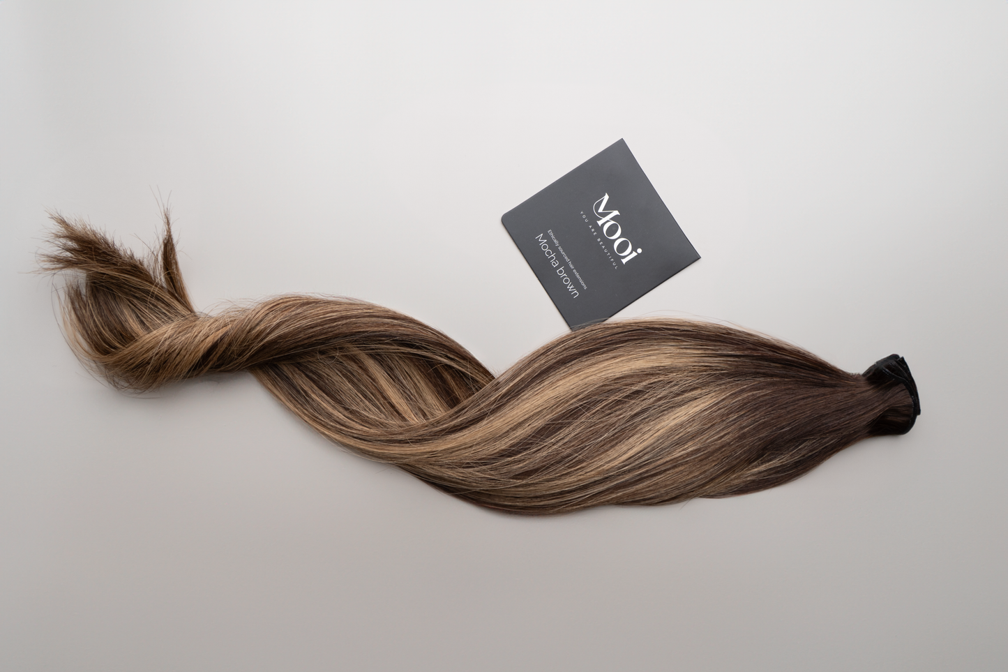 A twisted lock of brown and blonde hair, secured at the top, rests on a light surface. Next to it is a black card featuring the brand "Mooi Hair" and some text, suggesting their Seamless Weft | 50cm/20 inch hair extensions—a product designed to offer a flawless blend and customized fit.