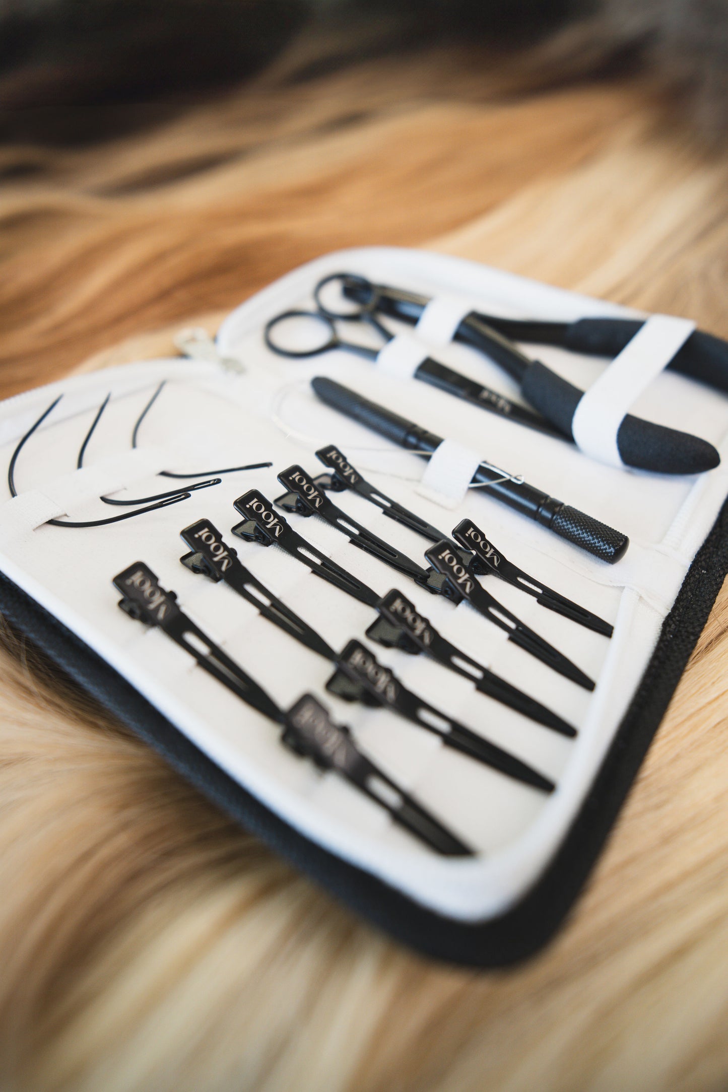 For the Mooi Hair extensions workshop, a close-up reveals an open hair styling kit designed for seamless weft and silk weft hands-on training. The black and white kit includes neatly organized scissors, hair clips, and brushes. It sits on a surface with flowing blonde and brown hair in the background.
