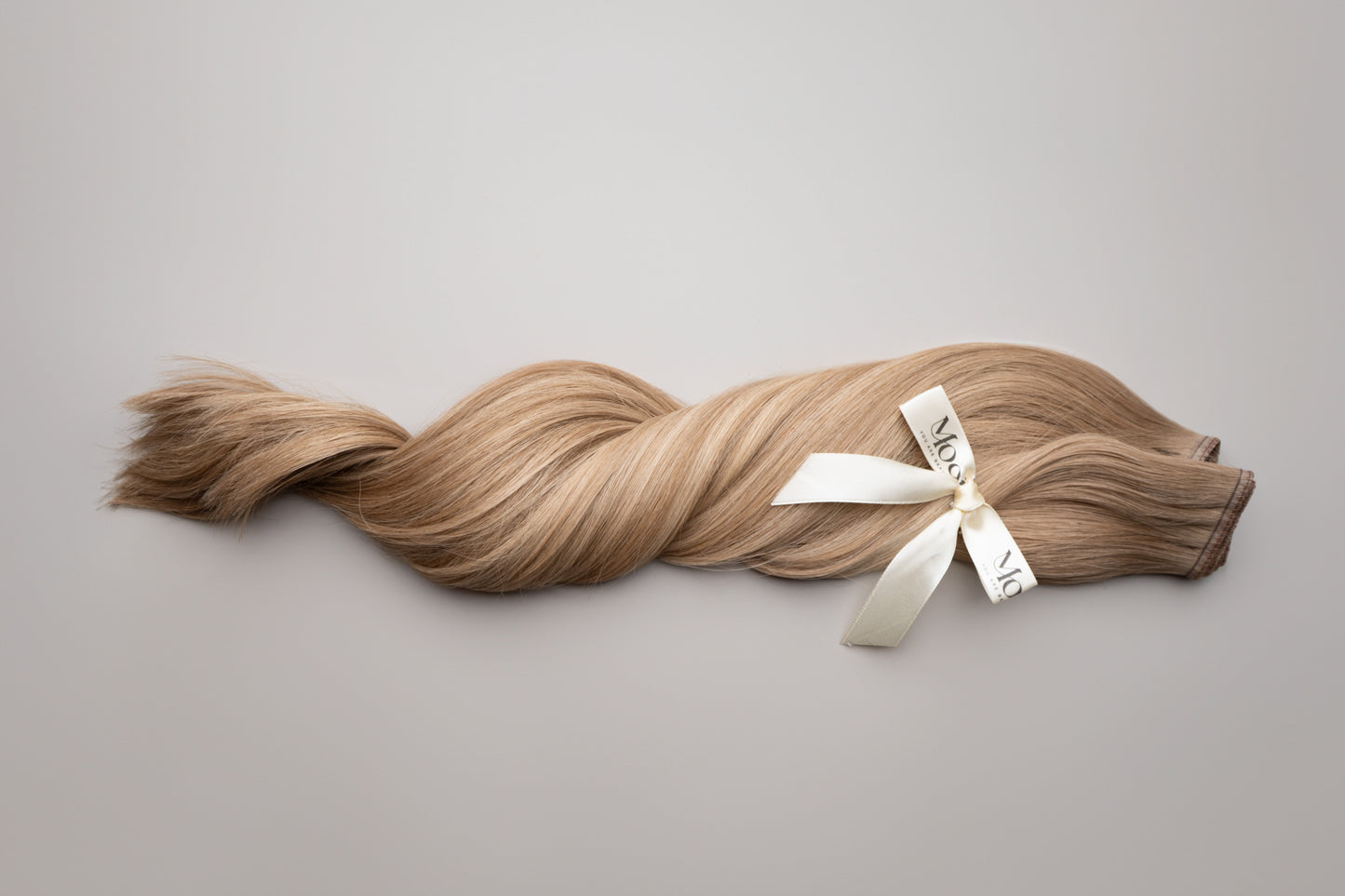 A luxurious braid of silky, light brown hair with a smooth texture is tied with a white ribbon emblazoned with "Mooi Hair," laid elegantly on a neutral background. The Silk Weft | 50cm/20 inch product ensures versatile styling while maintaining a premium feel with 100% cuticle-correct strands.