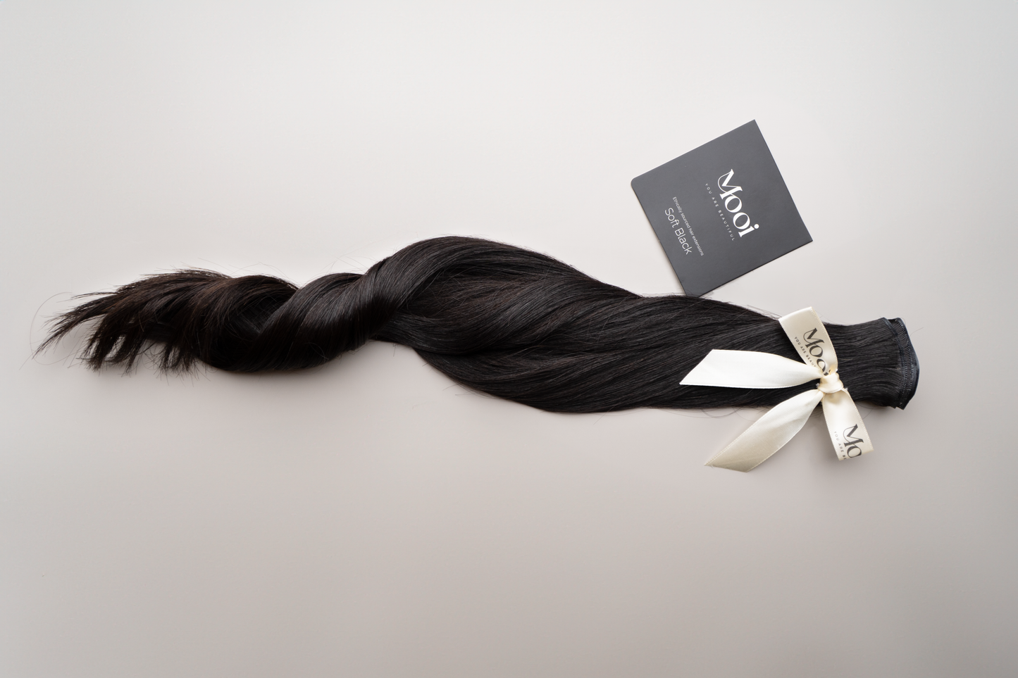 A bundle of the Seamless Weft | 50cm/20 inch from Mooi Hair, featuring long, dark brown hybrid wefts, is secured with a cream-colored ribbon. A black tag with white text accompanies the bundle, all displayed on a light gray background to highlight these seamless hair extensions designed for a flawless look.