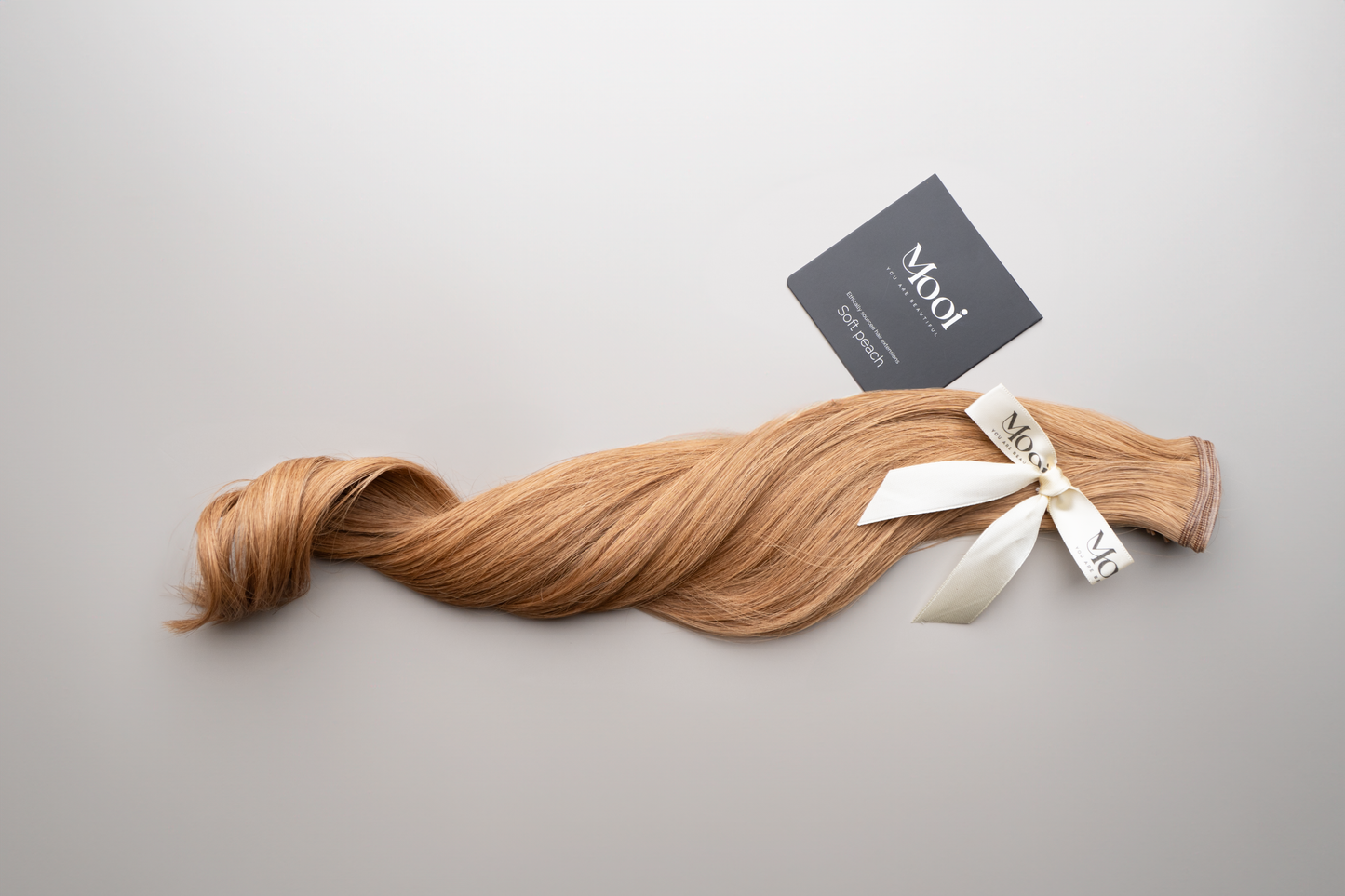 A long strand of wavy blonde hair, tied with a white ribbon and labeled "Mooi Hair," along with a black card, showcasing the Seamless Weft | 50cm/20 inch hair extensions, is laid on a light grey background.