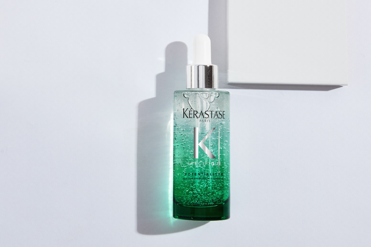 A bottle of Specifique Potentialiste Serum 90ml from Kerastase rests on a white surface. Its sleek design showcases a green gel with a dropper lid, casting an elegant shadow. Nearby, a partially visible white box hints at its luxurious nature.