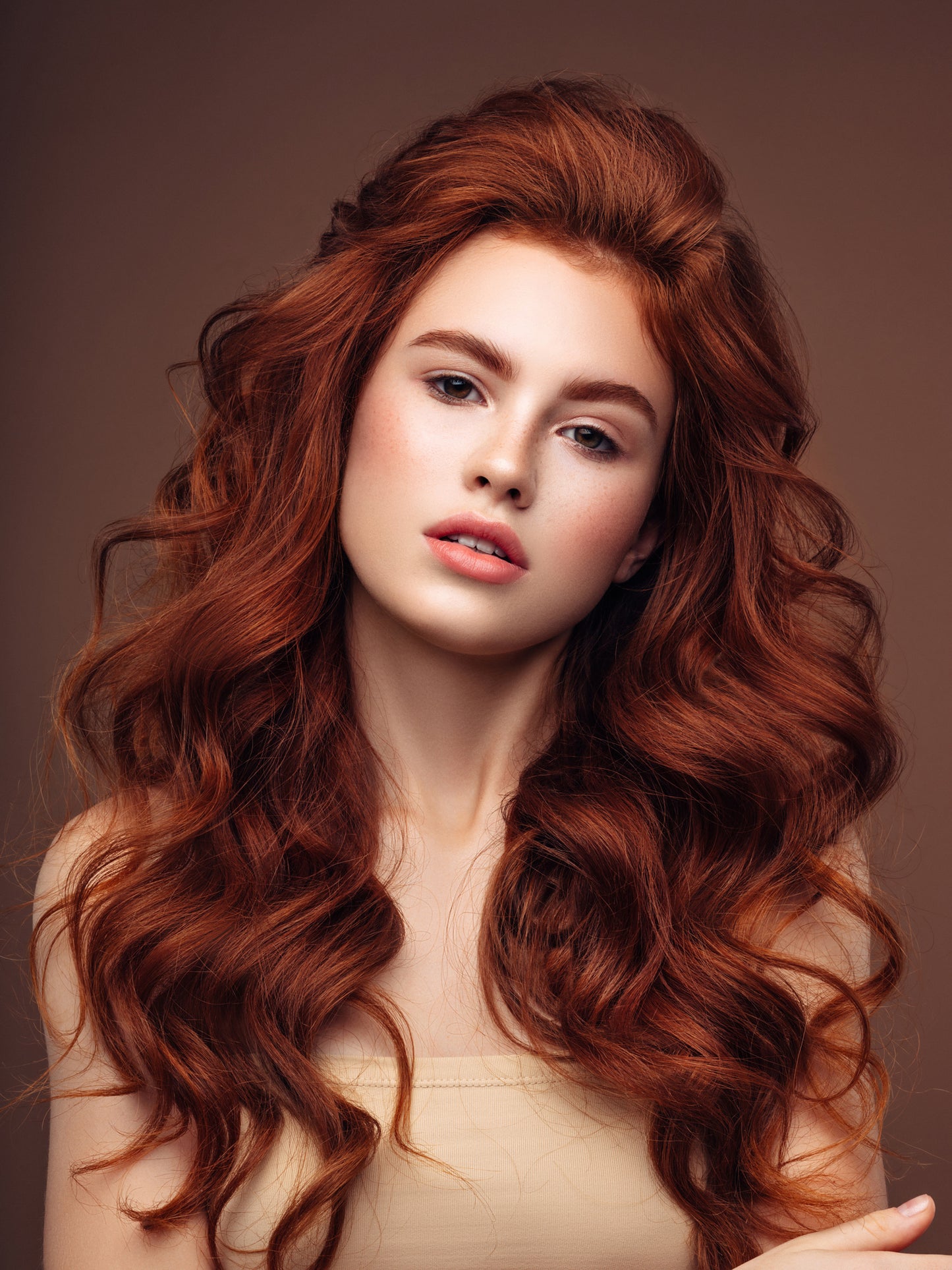 A young woman with long, wavy red hair styled using techniques from the Bonds & Weft Hands On Masterclass by Mooi Hair poses against a brown background. She has fair skin and is wearing a strapless beige top. Her hair falls elegantly around her shoulders, and her expression is calm and composed.