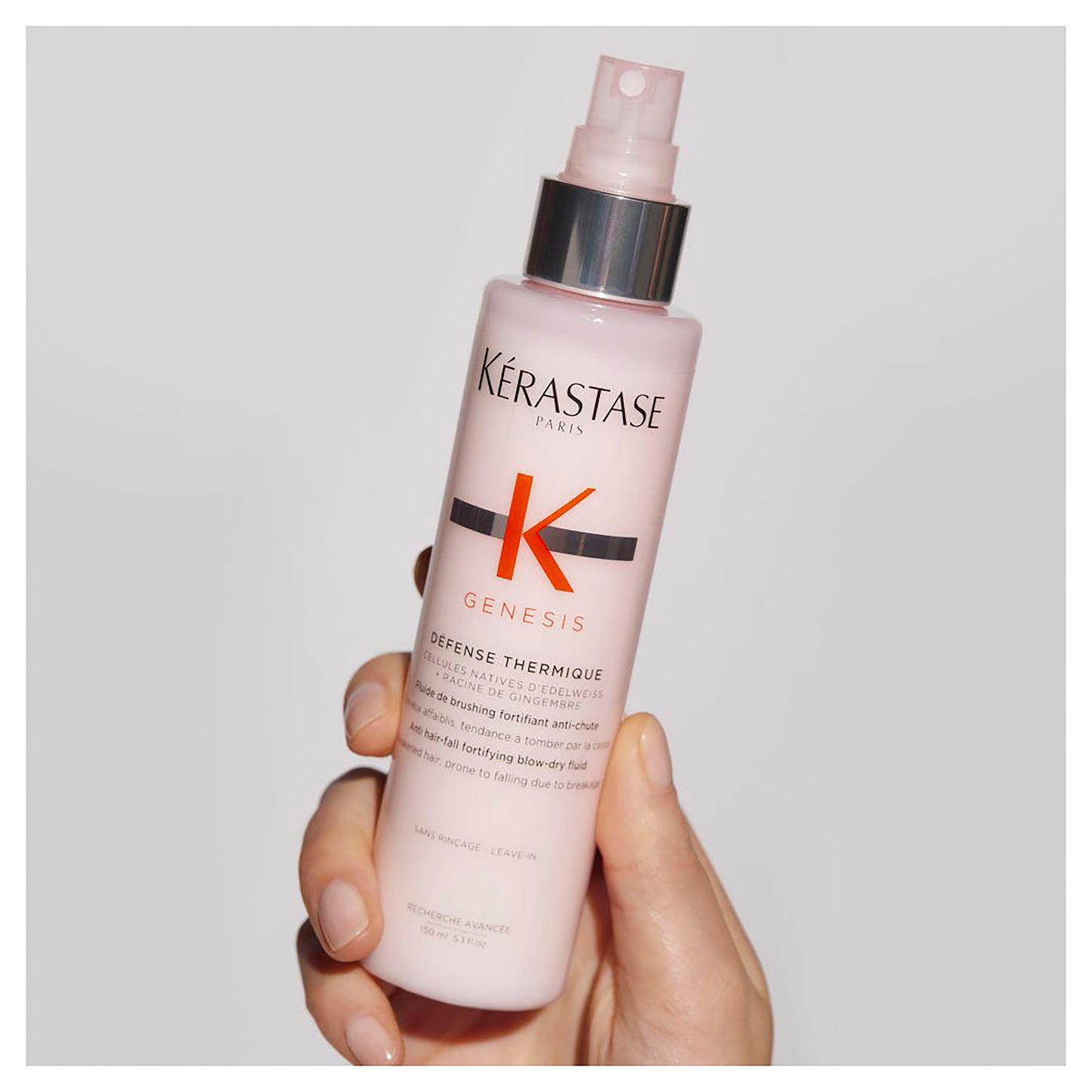 A hand holds a pink spray bottle of Kérastase Genesis Defense Thermique 150ml, highlighting the heat-protective blow-dry fluid. The bottle comes with a silver pump and is adorned with an eye-catching orange "K" logo, enhancing its striking visual appeal.