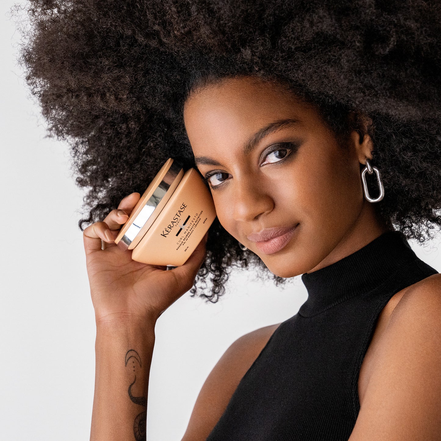 A person with curly hair, adorned in a sleeveless black top and hoop earrings, holds a Curl Manifesto Masque Beurre Haute Nutrition 200ml by Kérastase next to their face. A tattoo embellishes their arm as they gaze directly at the camera, exuding style and confidence.